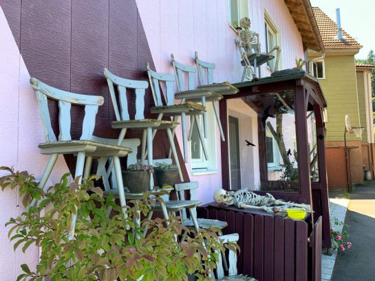 Ein Haus, teils rosa gestrichen. Holzstühle sind an der Fassade angebracht. Sie bilden eine Treppe. Vor der Haustür liegen Plastikskelette. 