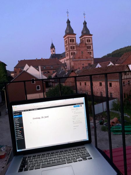 Balkon. Vordergrund: Laptop auf dem Schoss, Backend des Blogs. Im Hintergrund zwei Türme einer Kirche, Altstadt. 