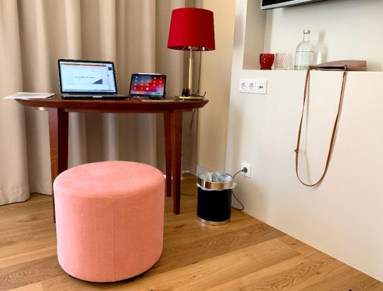 Hotelzimer: Tisch mit MacBook und iPad. Eine Lampe mit rotem Schirm steht daneben. Davor ein rosa Hocker. 