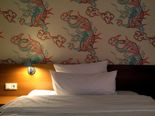 Hotelbett mit Nachttischlampe und Tapete: weiß mit blau-roten Drachen