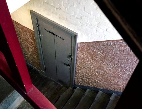 Blick durch das Treppengeländer nach unten. Auf 12 Uhr eine Tür mit der Aufschrift "Hoteltechnik", Industriecharme. 
