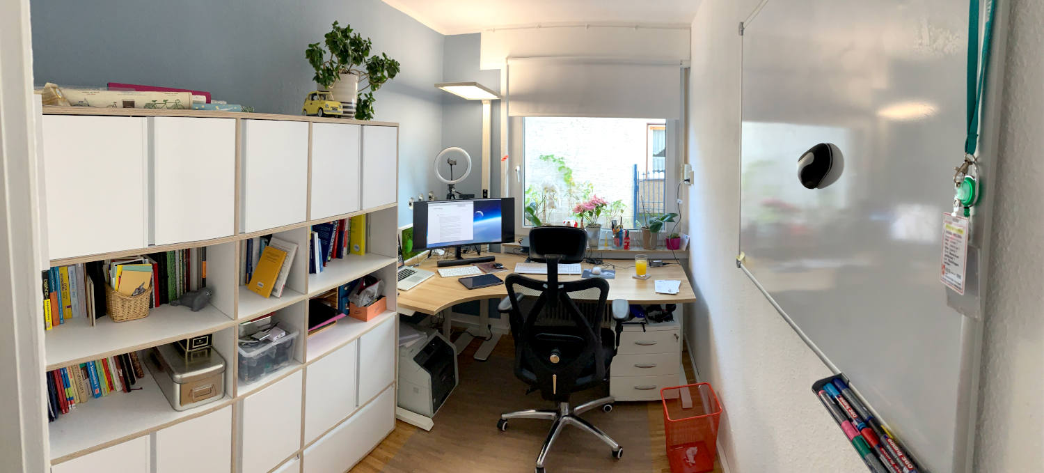 Panoramaaufnahme eines kleinen Arbeitszimmer: Schreibtisch vor dem Fenster, links eine Regal-Schrankwand, rechts ein Whiteboard