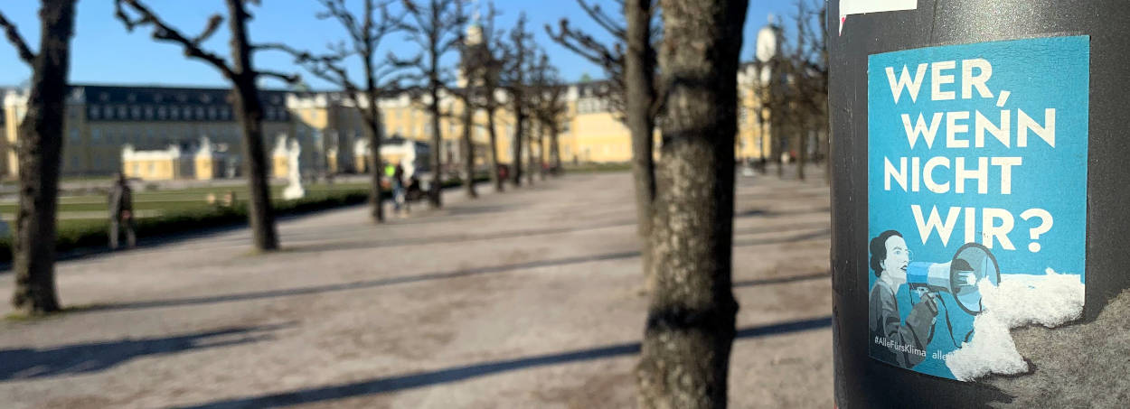 Park, im Hintergrund das Karlsruher Schloss, im Vorderung ein Aufkleber "Wer, wenn nicht wir?"