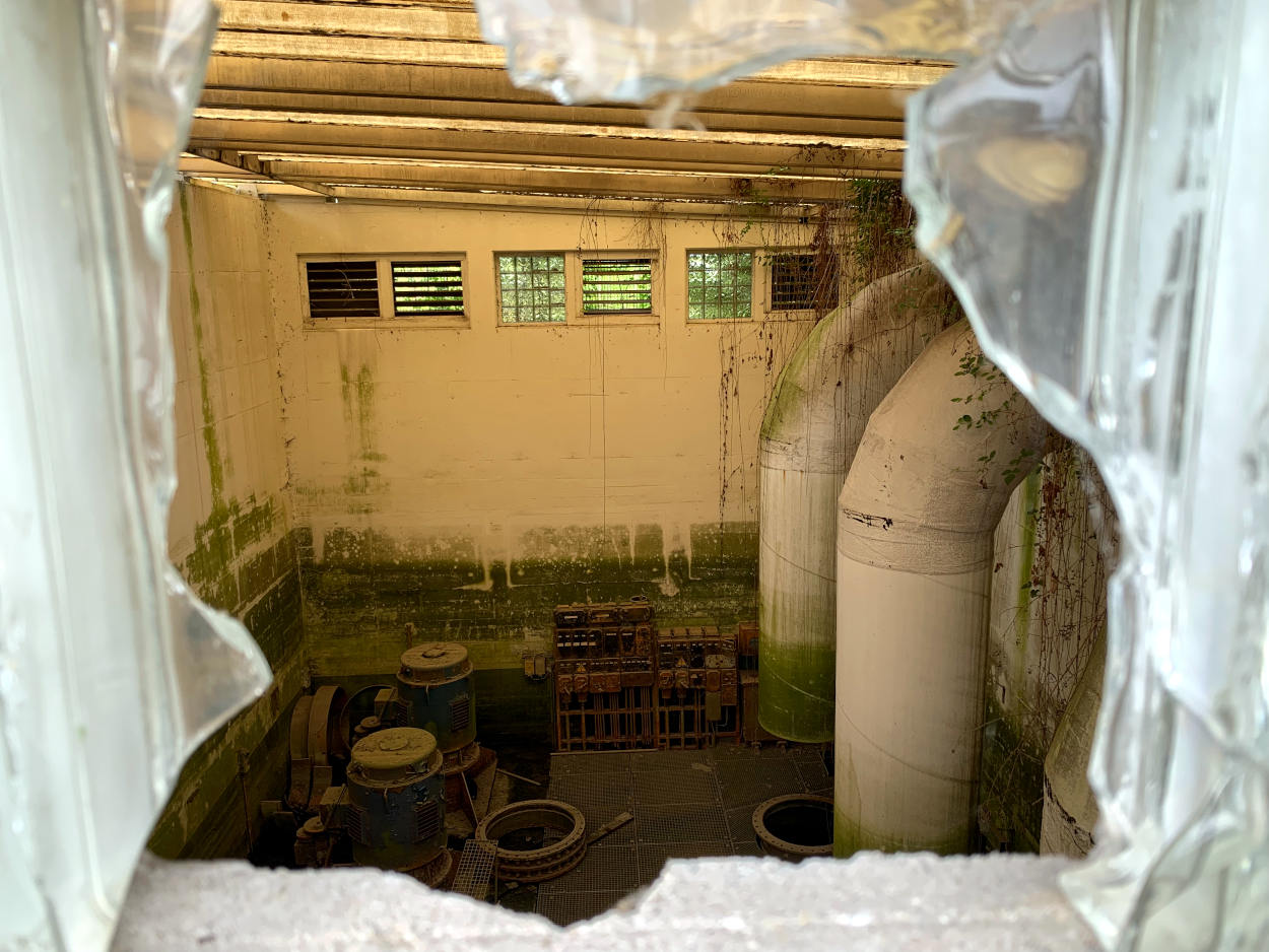 Blick durch ein zerschlagens Fenster in ein altes Wasserwerk: Rohre sind zu sehen. 