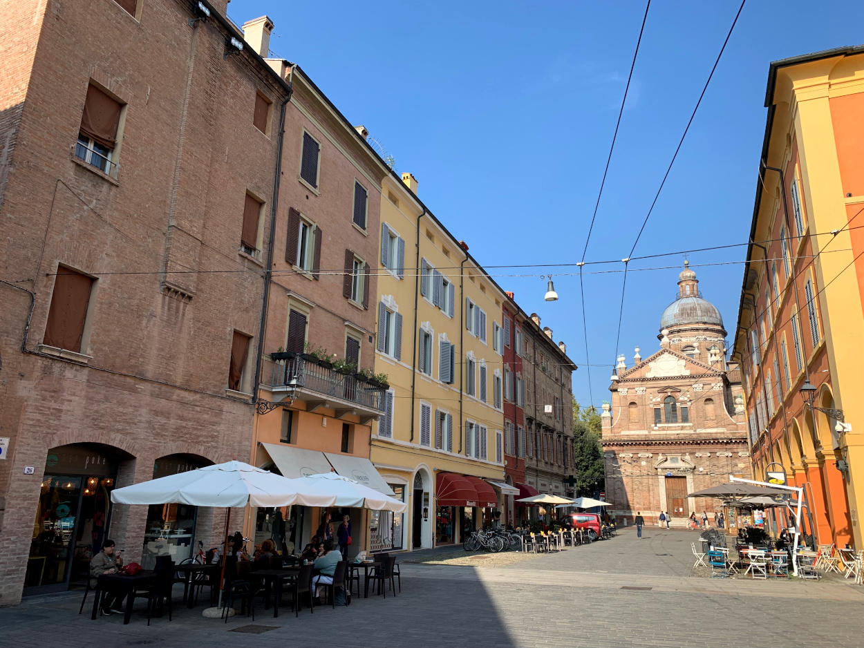 Platz in Modena, vor Kopf eine Backsteinkirche, auf dem Platz Tische und Menschen in der Mittagspause