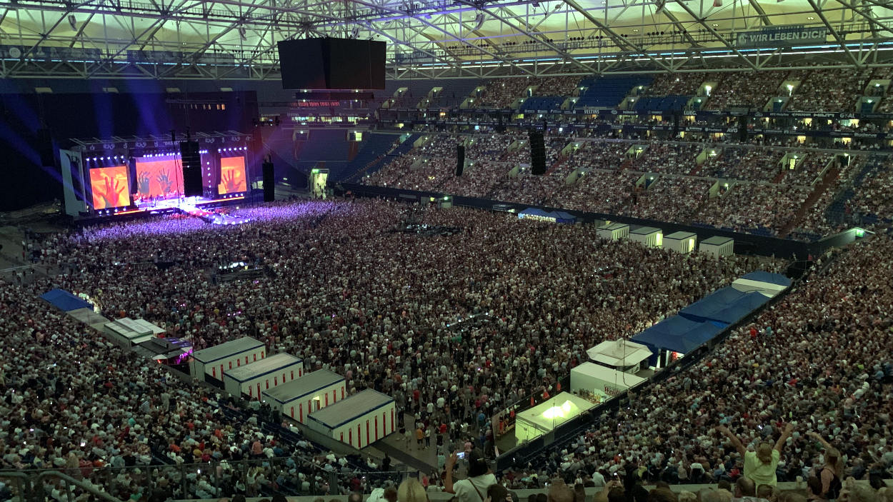 Arena auf Schalke, proppenvoll mit Menschen, auch im Innenraum, im Hintergrund eine Bühne. Das Konzert ist im Gange. 