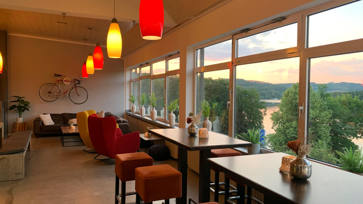 Restaurant: An der Wand ein Fahrrad, gelbe und rote Lampen, eine breite Fensterfront mit Blick auf den Biggesee im Abendrot