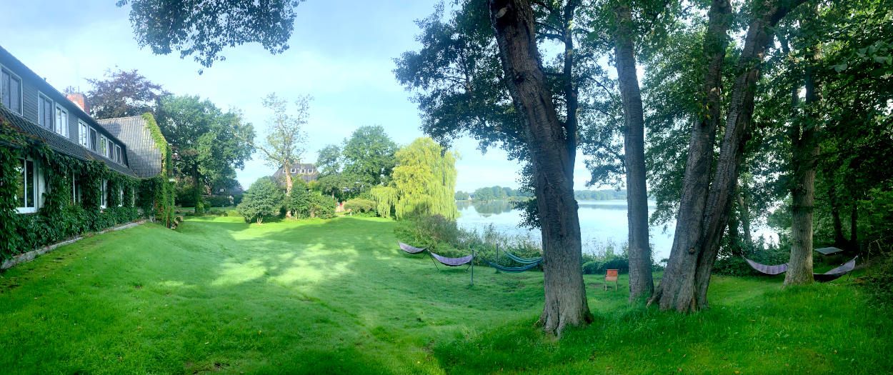Panorama-Aufnahme einer Wiese, im Hintergrund der See. Auf der Wiese stehen Hängematten