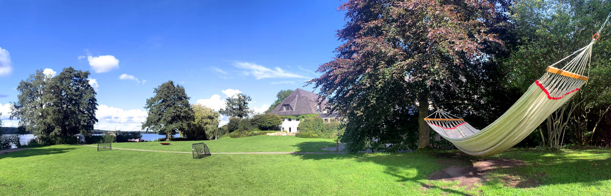 Panoramaaufnahme einer Wiese, im Hintergrund ein Haus mit Reetdach, am Rande eine riesige Hängematte in den Bäumen
