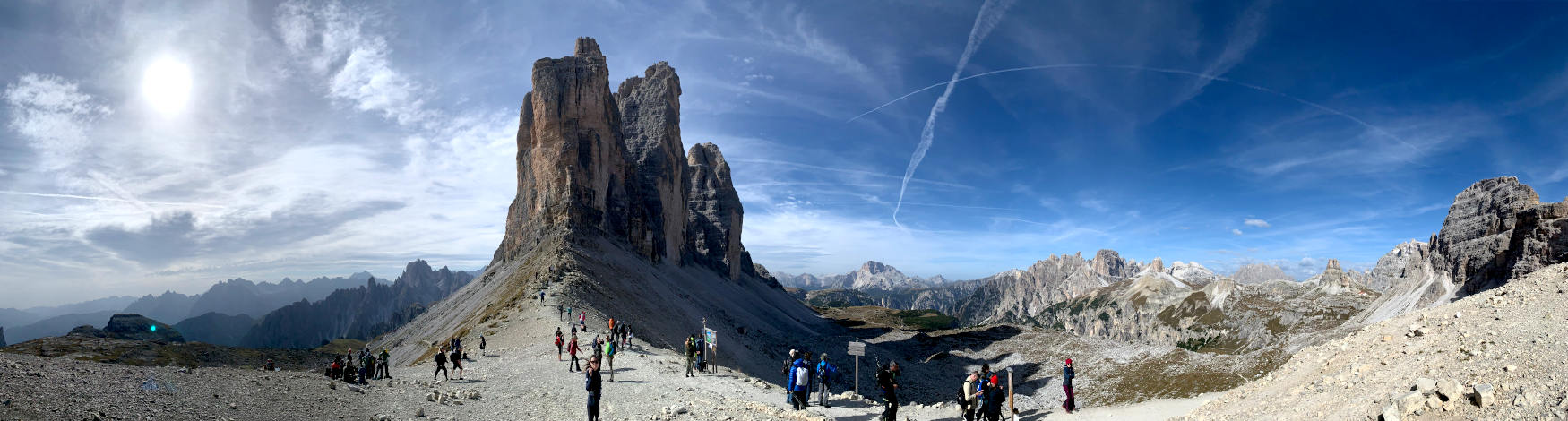 Panoramabild, in der Mitte die drei Zinnen, an ihrem Fuß Menschen. Im Hintergrund weitere Berge. 