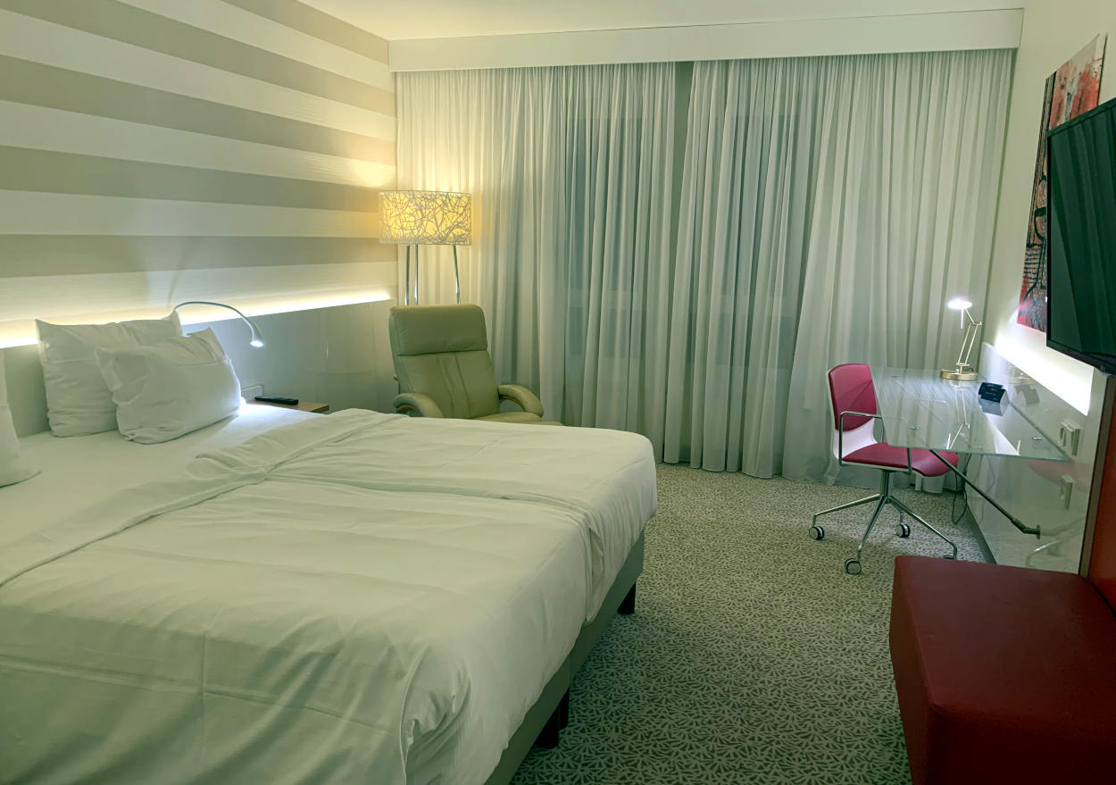 Hotelzimmer: Links ein Doppelbett, die Tapete dahinter gestreift. Vor dem Fenster (geradeaus) eine weiße, blickdichte Gardine. Links ein Glasschreibtisch mit rotem Stuhl davor und ein Fernseher an der Wand. Die Atmosphäre ist kühl.