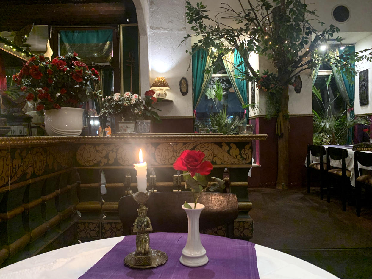 Blick in ein überladen dekoriertes Restaurant. Zwischen zwei Fenstern steht ein künstlicher Baum, es gibt Vorhänge, Palmen und rot blühende Blumen, auf dem Tisch eine Kerze und eine Rose, grün-gold-verzierte Ballustraden. 