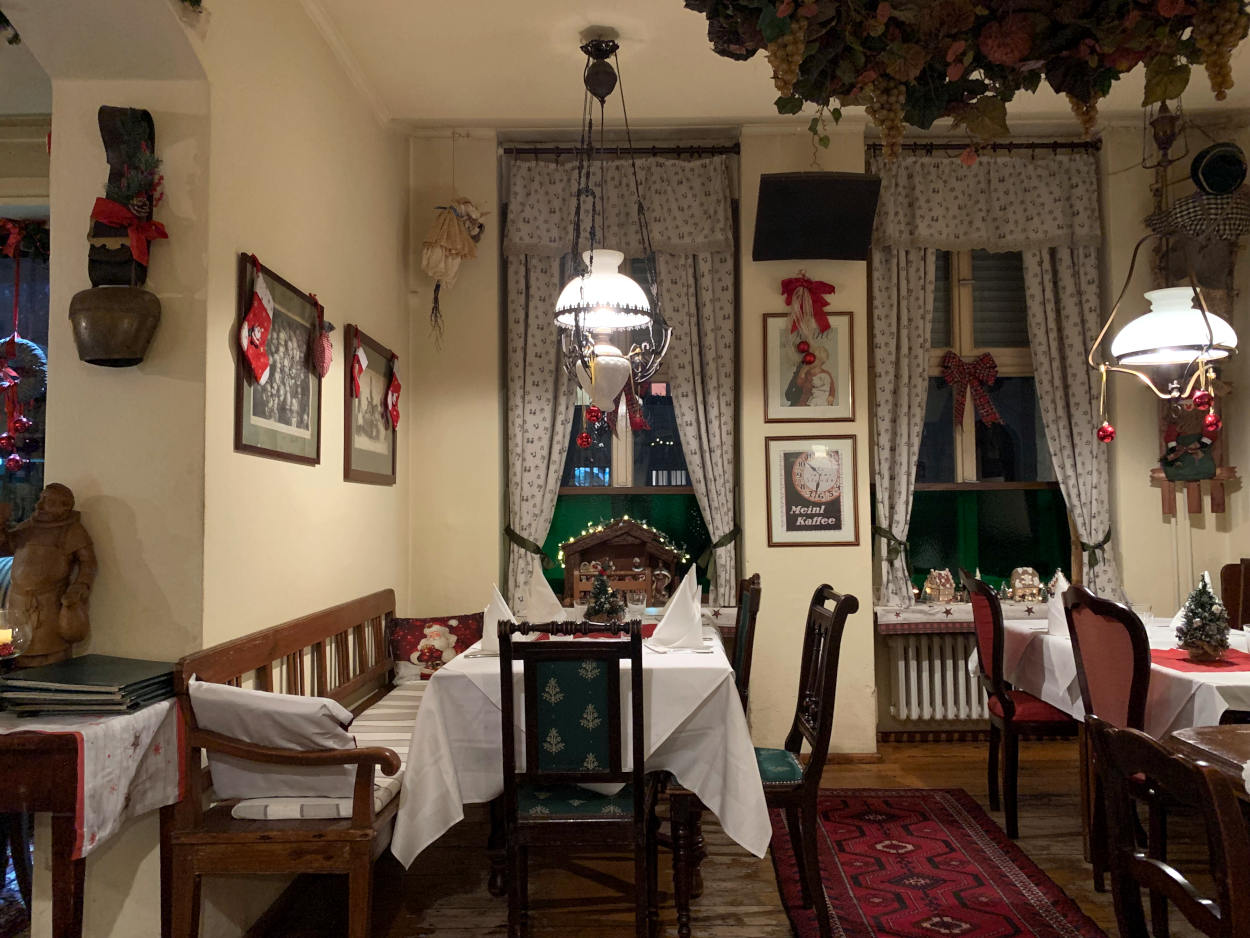 Restaurant, das aussieht wie eine gute Stube: Hängegardinen, traditionelle Lampen, Stühle mit Polster und Bänke zum Sitzen. An den Wänden eine Kuhglocke und Bilder