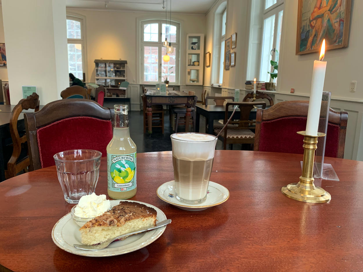 Antikes Café-Ambiente, auf dem Tisch eine Kerze, ein Milchkaffee und Kuchen, außerdem eine Limonade