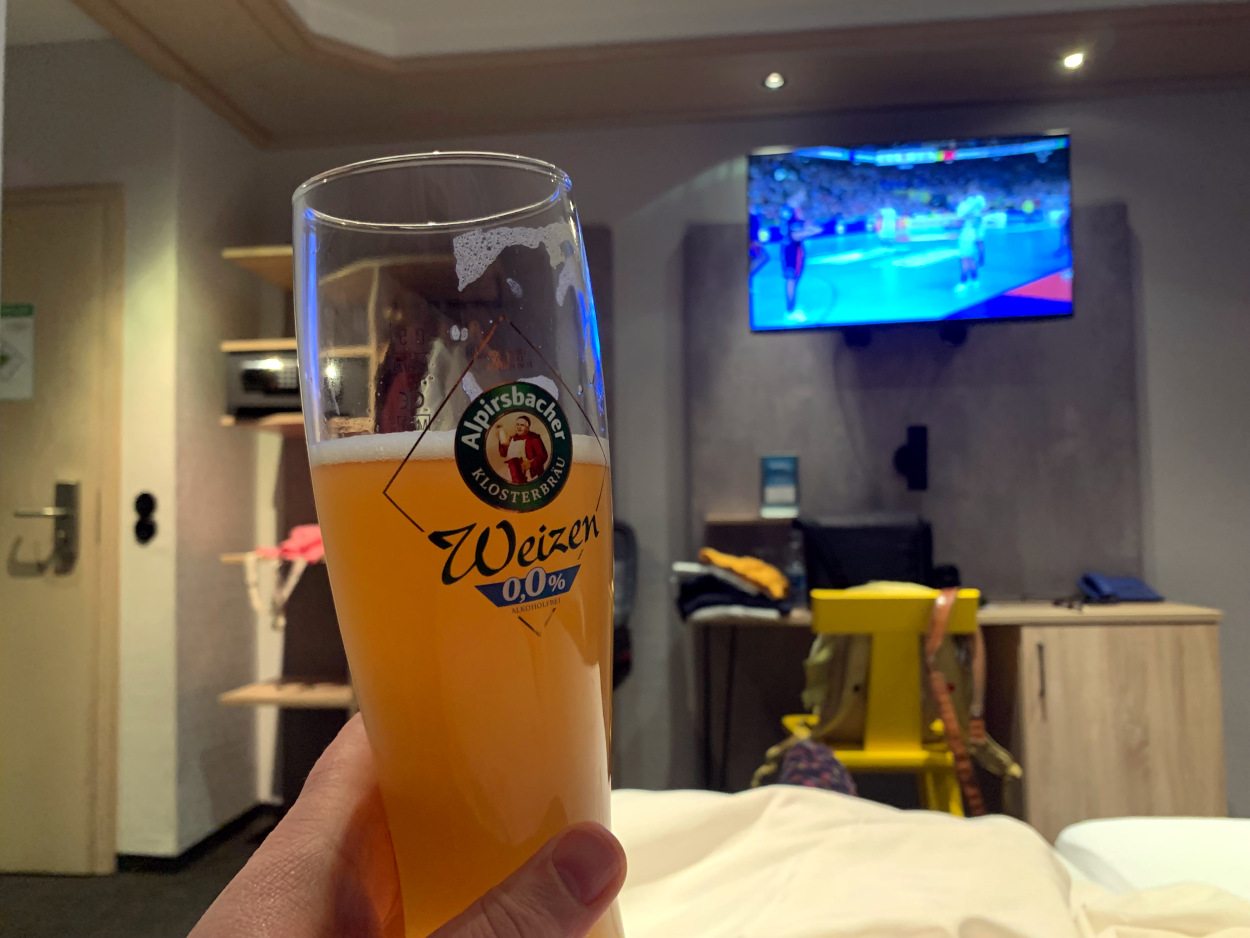 Foto vom Hotelbett aus: Im Vordergrund ein alkoholfreies Weizenbier in einer Hand, im Hintergrund ein Fernseher an der Wand mit Handball