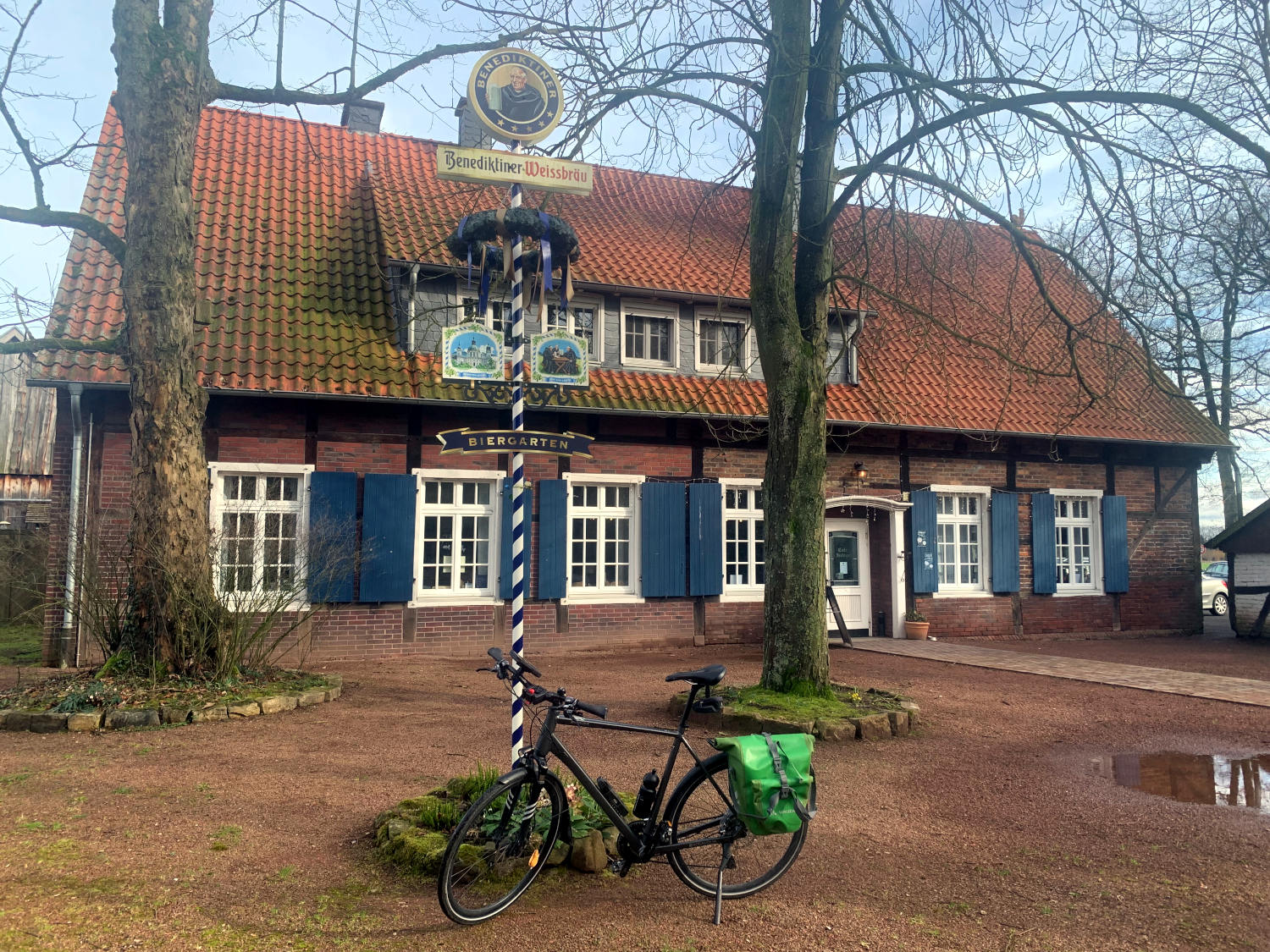 Münsterländer Landhaus, Backstein mit blauen Fensterläden. Davor ein Maibaum und Bäume - und ein Fahrrad. 